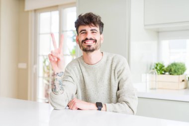 Genç adam rahat kazak beyaz masanın üzerinde oturan giyen kamera parmaklar zafer işareti yapıyor gösteren bakıyor. İki numara.