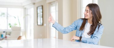 Evde beyaz masada oturan güzel genç kadının geniş açı resim gururlu görünümlü, yan hareket başparmak kadar yapıyor gülümseyen