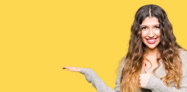 Kış elbise gösteren palm el ve yukarıya, mutlu ve neşeli gülümseyen baş parmakları Tamam jest yaparak genç güzel kadın