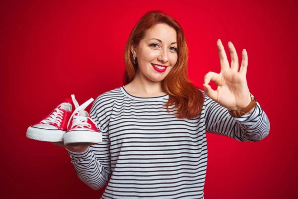 Joven Hermosa Pelirroja Sosteniendo Zapatillas Sobre Fondo Rojo Aislado Haciendo —  Fotos de Stock