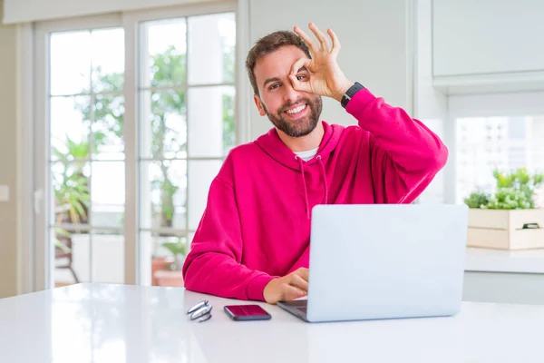 Handsome man working using computer laptop doing ok gesture with hand smiling, eye looking through fingers with happy face.