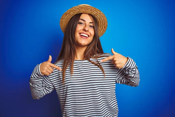Young beautiful woman wearing navy striped t-shirt and hat over isolated blue background looking confident with smile on face, pointing oneself with fingers proud and happy.