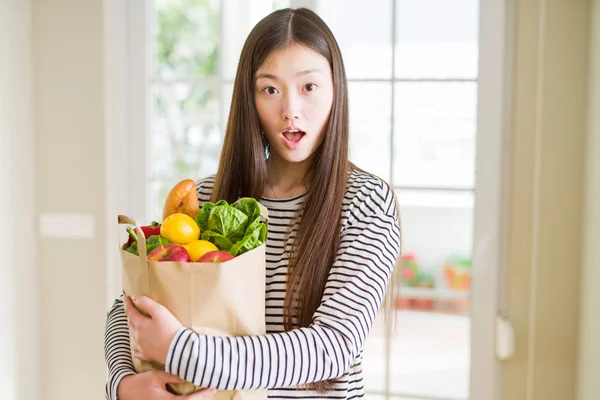 Beautiful Asian woman holding paper bag of fresh groceries scared in shock with a surprise face, afraid and excited with fear expression