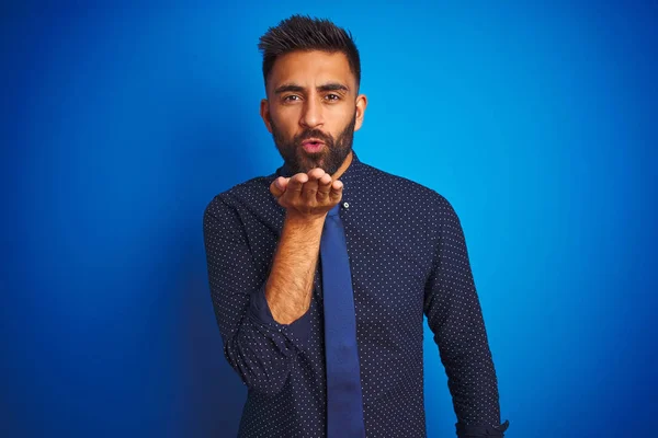 年轻的印度商人穿着优雅的衬衫和领带站在孤立的蓝色背景上看着相机吹一个吻与手在空中可爱和性感 爱表达 — 图库照片