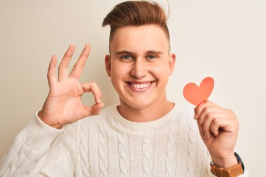 Genç yakışıklı romantik adam parmaklar, mükemmel sembolü ile ok işareti yapıyor izole beyaz arka plan üzerinde duran kağıt kalpleri tutan