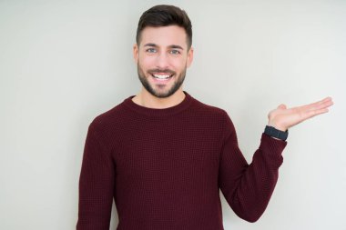 Genç yakışıklı adam izole arka plan üzerinde bir kazak giyen neşeli sunan gülümseyerek ve kameraya bakarak avuç içi ile işaret.