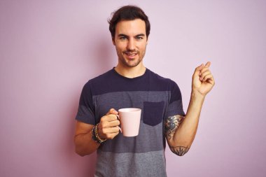 Dövme ile genç yakışıklı adam izole pembe arka plan üzerinde kahve içme bardağı yan el ve parmak ile işaret çok mutlu