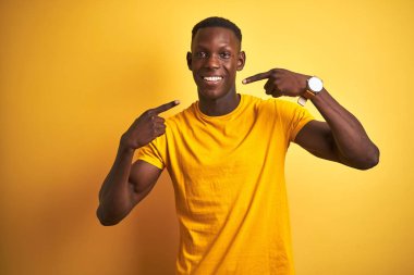 İzole sarı arka plan üzerinde ayakta rahat t-shirt giyen genç Afrikalı Amerikalı adam neşeli gösteren gülümseyen ve parmaklar diş ve ağız ile işaret. Diş Sağlığı konsepti.