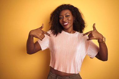 Izole sarı arka plan üzerinde duran pembe t-shirt giyen Afro-Amerikan kadın yüzünde gülümseme ile emin görünümlü, parmakları gururlu ve mutlu kendini işaret.