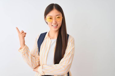 Yüz üzerinde büyük bir gülümseme ile izole beyaz arka plan üzerinde gözlük ve sırt çantası giyen genç Çinli öğrenci kadın, kamera bakarak yan el ve parmak ile işaret.