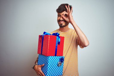 Dövme tutan Doğum günü hediyeleri ile genç adam mutlu yüz gözleri parmaklarıyla görünümlü el ile ok işareti yapıyor gülümseyen izole beyaz arka plan üzerinde ayakta