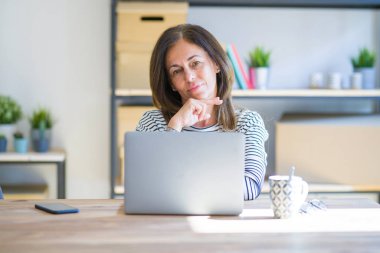 Orta yaş kıdemli kadın evde masada oturan bilgisayar laptop kullanarak kamera emin görünümlü karşı karşıya silah ve el çene üzerinde yükseltilmiş ile gülümseme ile çalışıyor. Pozitif düşünme.