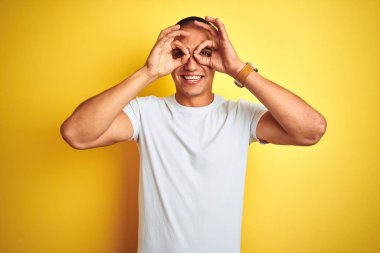 Sarı izole arka plan üzerinde rahat beyaz t-shirt giyen genç beyaz adam dürbün yapışması gibi ok jest yapıyor, gözler parmaklarla bakıyor. Çılgın ifade.