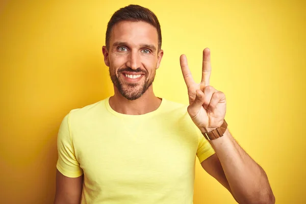 黄色の孤立した背景の上にカジュアルな黄色のTシャツを着た若いハンサムな男は 自信を持って幸せに微笑みながら 指ナンバー2で見せ 指を上げる — ストック写真