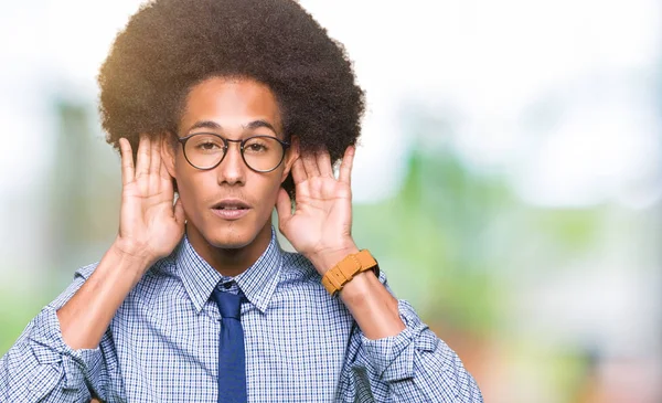Young african american business man with afro hair wearing glasses Trying to hear both hands on ear gesture, curious for gossip. Hearing problem, deaf