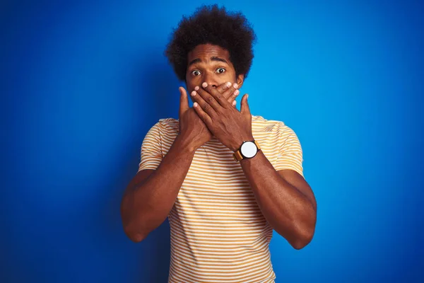 American man with afro hair wearing striped yellow t-shirt over isolated blue background shocked covering mouth with hands for mistake. Secret concept.