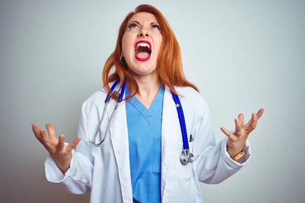 白い孤立した背景の上に聴診器を使用して若い赤毛の医師の女性は狂気と狂った叫び声と積極的な表情と腕を上げて叫ぶ 欲求不満の概念 — ストック写真