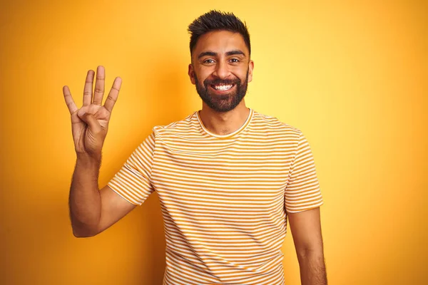 孤立した黄色の背景の上に立つTシャツを着た若いインド人男性は 自信を持って幸せに微笑みながら 4番の指を見せ 指を上げている — ストック写真