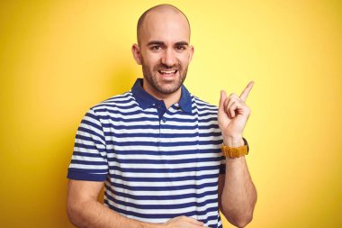 Yüzünde büyük bir gülümseme ile sarı izole arka plan üzerinde rahat çizgili mavi t-shirt giyen sakal ile genç kel adam, kamera bakarak yan el ve parmak ile işaret.