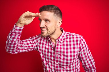 Kırmızı izole arka plan üzerinde genç yakışıklı adam çok mutlu ve gülümsüyor uzağa baş el ile bakıyor. Arama konsepti.