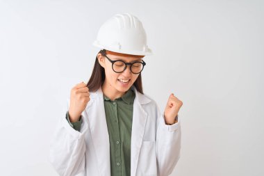 Genç Çinli mühendis kadın izole beyaz arka plan üzerinde ceket kask gözlükleri giyen çok mutlu ve heyecanlı silah kaldırdı, gülümseyen ve başarı için çığlık ile kazanan jest yapıyor. Kutlama konsepti.