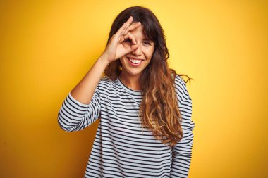 Yelllow izole arka plan üzerinde ayakta çizgili t-shirt giyen genç güzel kadın el gülümseyen ile ok jest yapıyor, mutlu yüz ile parmaklar görünümlü göz.
