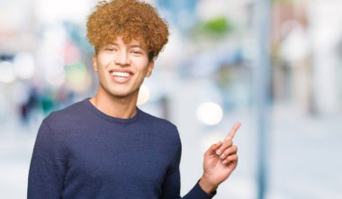 Yüzünde kocaman bir gülümseme ile afro saçlı genç yakışıklı adam kameraya bakarak yan el ve parmak ile işaret.