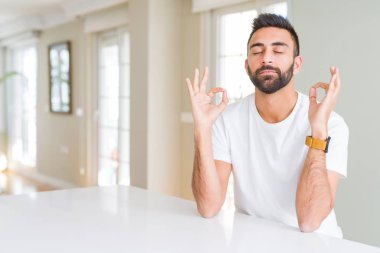 İspanyol yakışıklı casual beyaz t-shirt evde dinlenmek ve gülümseyen gözlerle parmak ile meditasyon jest yapıyor kapalı. Yoga kavramı.