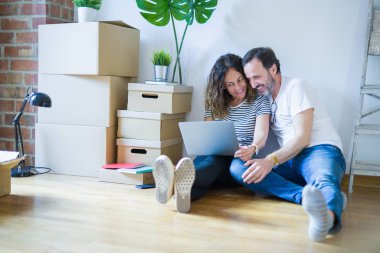 Orta yaş üst düzey romantik çift aşk etrafında kutuları ile daire katında oturan ve bilgisayar laptop kullanarak yeni bir eve hareket için mutlu gülümseyen