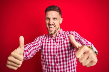 Kırmızı izole arka plan üzerinde genç yakışıklı adam el ile olumlu jest yapıyor onaylanır, gülümseyen ve başarı için mutlu kadar başparmak. Kazanan jest.