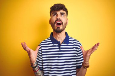 Dövme ile izole sarı arka plan üzerine çizgili polo ayakta giyen genç adam çılgın ve deli bağırma ve agresif ifade ve silah ile bağırdı kaldırdı. Hayal kırıklığı kavramı.