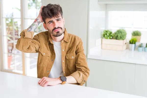 Young man wearing casual jacket sitting on white table confuse and wonder about question. Uncertain with doubt, thinking with hand on head. Pensive concept.