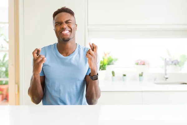 ハンサムなアフリカ系アメリカ人男性が自宅でカジュアルなTシャツを着て 希望と目を閉じて指を横切って微笑む 運と迷信的な概念 — ストック写真