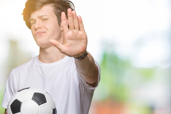 Young man holding soccer football ball over isolated background with open hand doing stop sign with serious and confident expression, defense gesture