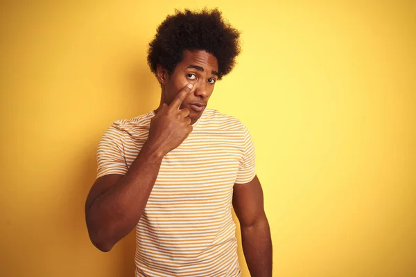 American man with afro hair wearing striped t-shirt standing over isolated yellow background Pointing to the eye watching you gesture, suspicious expression