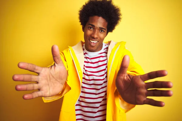 African american man with afro hair wearing rain coat standing over isolated yellow background looking at the camera smiling with open arms for hug. Cheerful expression embracing happiness.