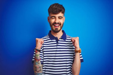 Çizgili polo giyen dövmeli genç adam izole mavi arka plan üzerinde duran kollarını kaldırdı ve gözleri gülümseyerek zafer kutlayan kapalı başarı için heyecanlı. Kazanan kavramı.