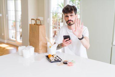 Genç adam eve teslim asya suşi yeme ve açık el ile akıllı telefon uygulaması kullanarak gıda sipariş ciddi ve kendinden emin ifade, savunma jesti ile işareti dur yapıyor