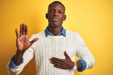 Izole sarı arka plan üzerinde denim gömlek ve beyaz kazak giyen Afro-Amerikan adam göğüs ve açık avuç içi el ile küfür, bir sadakat yemini yapma