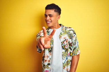 Tatilde yazlık çiçekli gömlek giyen brezilyalı genç adam izole sarı arka plan üzerinde gülümseyerek kameraya bakarak parmaklarını gösteren zafer işareti yapıyor. İki numara.