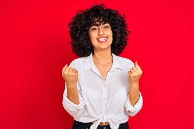 İzole kırmızı arka plan üzerinde beyaz rahat gömlek giyen kıvırcık saçlı genç Arap kadın şaşırmış ve kollarını kaldırdı ve gözleri açık başarı için şaşırttı. Kazanan kavramı.