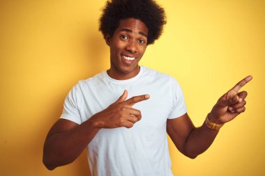 Beyaz saçlı afro saçlı Amerikalı adam izole sarı arka plan üzerinde duran beyaz t-shirt giyen gülümseyerek ve yan iki el ve parmakları ile işaret kamera bakıyor.