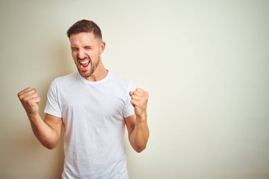 Gülümseyen ve başarı için çığlık izole arka plan çok mutlu ve heyecanlı kaldırdı, silah ile kazanan jest yapıyor üzerinde rahat beyaz t-shirt giyen genç yakışıklı adam. Kutlama kavramı.