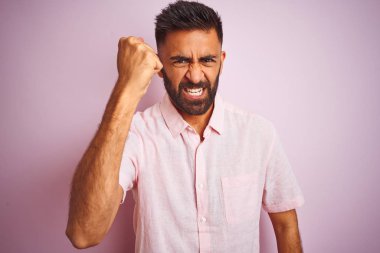Genç hintli adam izole pembe arka plan üzerinde duran rahat gömlek giyen kızgın ve deli yükselterek yumruk sinirli ve öfkeli öfke ile bağırırken. Öfke ve agresif kavram.