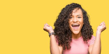 Kıvırcık saçlı pembe kutluyor t-shirt giyen genç güzel kadın şaşırttı ve başarı için kaldırdı kollarını açarak hayret ve gözleri açık. Kazanan kavramı.