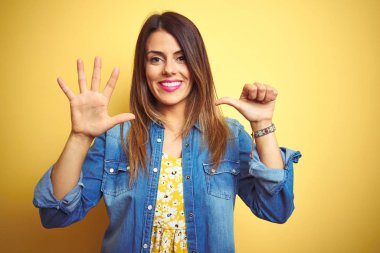 Genç güzel kadın sarı izole arka plan üzerinde duran gösteren ve parmakları ile işaret ederken emin ve mutlu gülümseyerek altı numara.