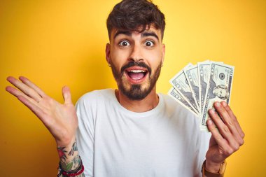 Dövme tutan dolar tutan genç adam izole sarı arka plan üzerinde duran çok mutlu ve heyecanlı, kazanan ifade büyük bir gülümseme ile çığlık zafer kutluyor ve ellerini kaldırdı