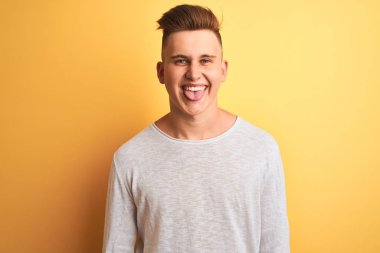 Genç yakışıklı adam beyaz rahat t-shirt giyen izole sarı arka plan üzerinde duran komik ifade ile mutlu dil yapışmasını. Duygu kavramı.