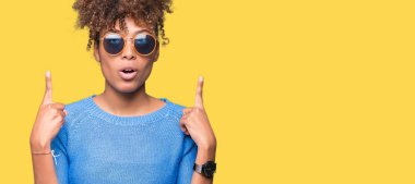 Güzel genç Afro-Amerikan kadın hayrete ve yukarıya bakıyor ve parmakları ile işaret ve silah kaldırdı sürpriz izole arka plan üzerinde güneş gözlüğü takıyor.