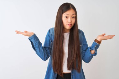 Denim gömlek giyen genç çinli kadın izole beyaz arka plan üzerinde duran habersiz ve kolları ve elleri ile karışık ifade kaldırdı. Şüphe kavramı.